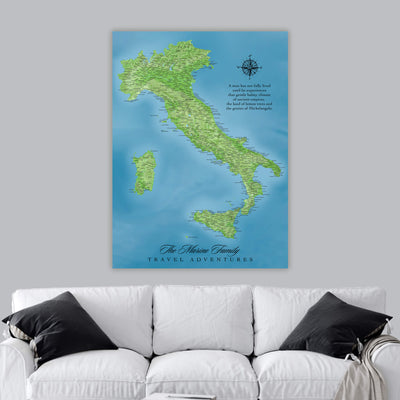 The image is a large poster of a map of Italy, with a compass rose and a quote about the joys of travel. The poster is displayed on a wall above a white couch with black pillows.