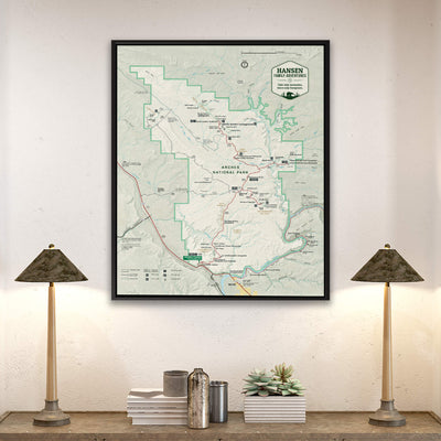 a framed map of the Hanging Forest National Park, with a lamp on a shelf below it and a stack of books on a table to the left.