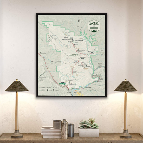 a framed map of the Hanging Forest National Park, with a lamp on a shelf below it and a stack of books on a table to the left.