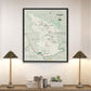 a framed map of the Hanging Forest National Park, with a lamp on a shelf below it and a stack of books on a table to the left.