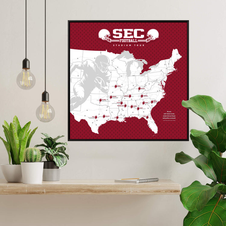 a framed map of the United States with football player silhouettes and the text "SEC Football Stadium Tour" printed on it. The map is displayed on a wall, and there are potted plants and a shelf with various items in the foreground.