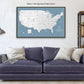 a large map of the United States with a blue background and white text, hanging on a wall above a blue couch with pillows.