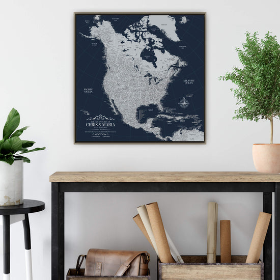 a large, detailed map of North America, with various regions and countries clearly labeled. The map is displayed on a wall, and there is a wooden table below it. On the table, there are some rolls of paper and a plant.