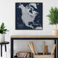 a large, detailed map of North America, with various regions and countries clearly labeled. The map is displayed on a wall, and there is a wooden table below it. On the table, there are some rolls of paper and a plant.