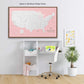 a workspace with a desk, chair, and various items such as a laptop, books, and a plant. Above the desk, there is a large map of the United States in a pink frame, and a smaller map of the world in a frame with a cartoon character on it.
