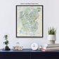 a framed map of the Rocky Mountain National Park region, with a black floater frame. Below the map, there is a glass dome containing a plant, a white clock, and a stack of books.