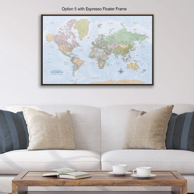 a large world map displayed on a wall above a white couch with two pillows. The map is framed in an espresso float frame, and there is a wooden coffee table in front of the couch with two white cups on it.