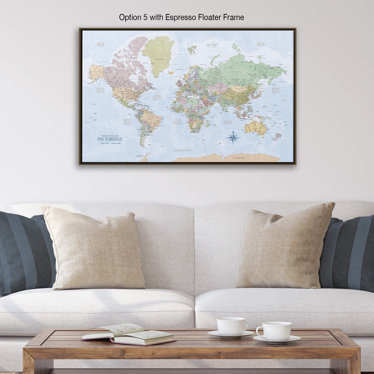 a large world map displayed on a wall above a white couch with two pillows. The map is framed in an espresso float frame, and there is a wooden coffee table in front of the couch with two white cups on it.