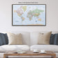 a large world map displayed on a wall above a white couch with two pillows. The map is framed in an espresso float frame, and there is a wooden coffee table in front of the couch with two white cups on it.