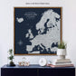 a large map of Europe with a brown frame, displayed on a wall above a wooden shelf. The map is surrounded by various decorative items, including a glass dome with a plant, a stack of books, and a white clock.