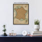 a framed map of France hanging on a wall, with a glass dome containing a plant and a stack of books below it.